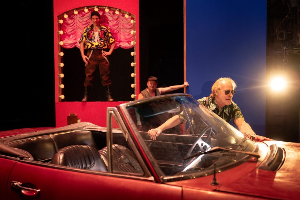 Ein Schauspieler steht auf der Bühne neben einem roten Cabrio.