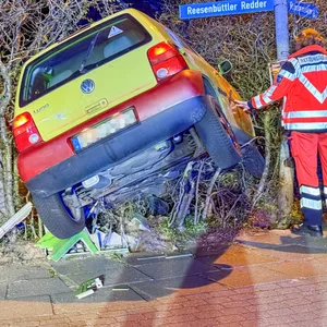 Der gelb/rote VW liegt in einer Hecke. Einsatzkräfte stehen daneben.