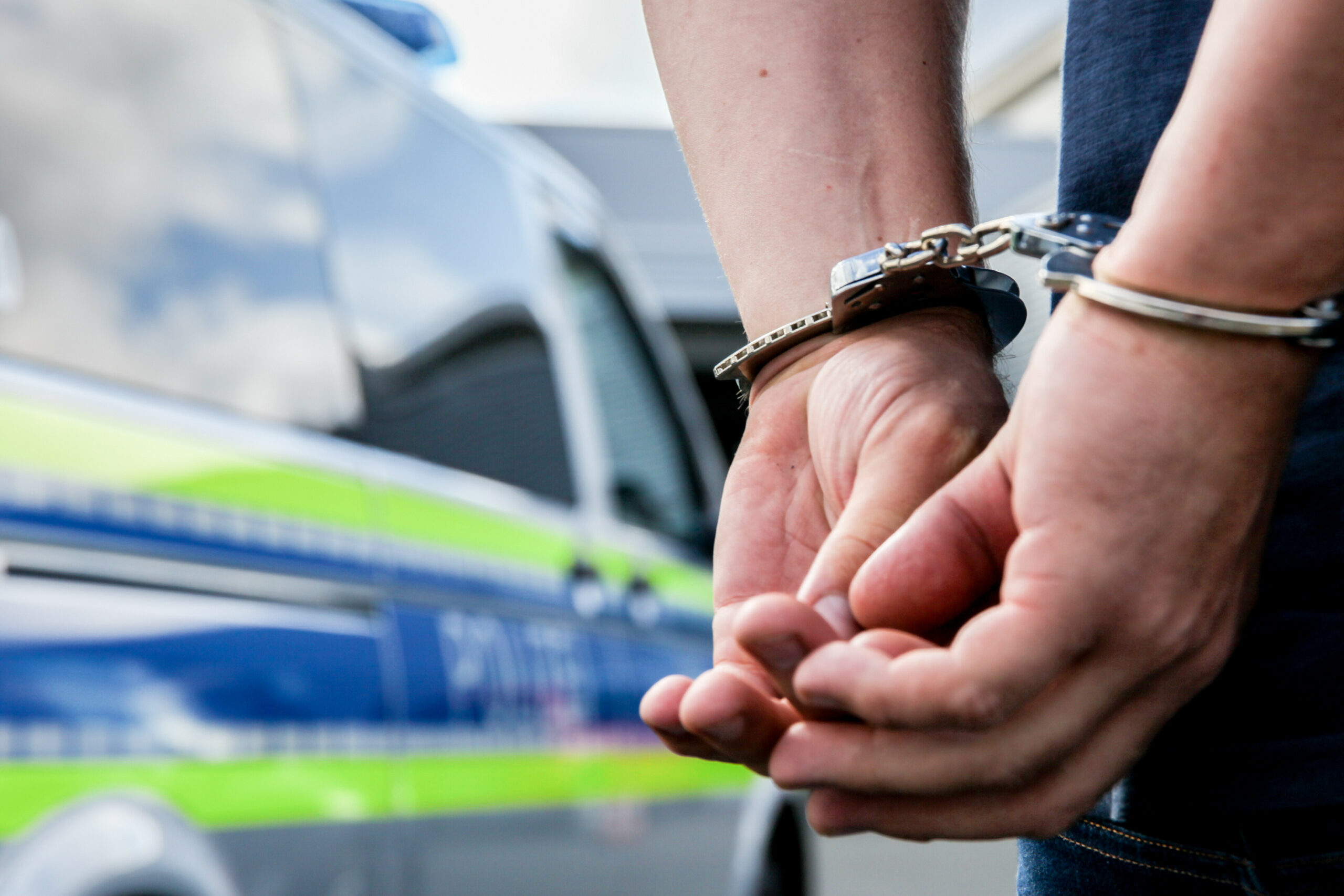 Eine Person steht in Handschellen gefesselt neben einem Polizeifahrzeug.