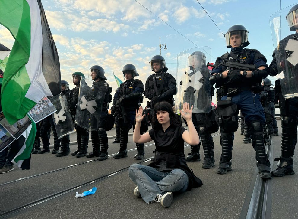 Eine Frau sitzt vor einer Reihe von Polizisten und hält ihre Hände hoch, auf denen „Land back“ (Land zurück) geschrieben steht.