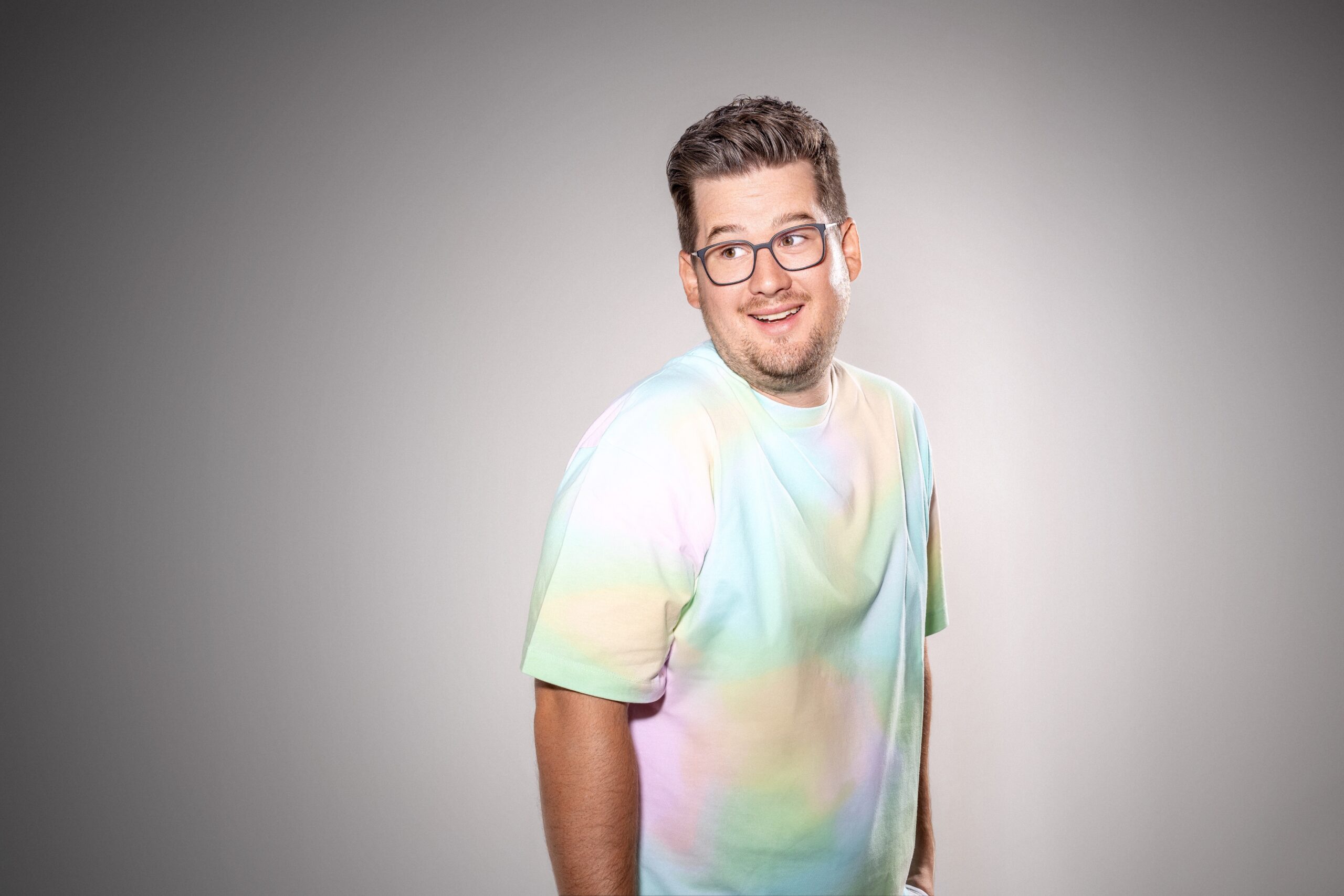 Er steht im T-Shirt vor einer Wand und lächelt verlegen
