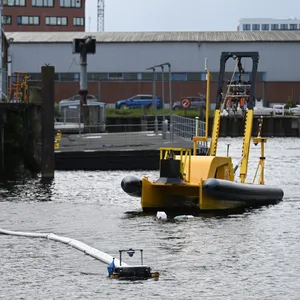Zwei schwimmende Roboter sammeln bei einem Pressetermin in Hamburg-Harburg Müll auf dem Wasser ein, im Hintergrund fährt ein Schiff, dass den Müll aufsammeln soll.
