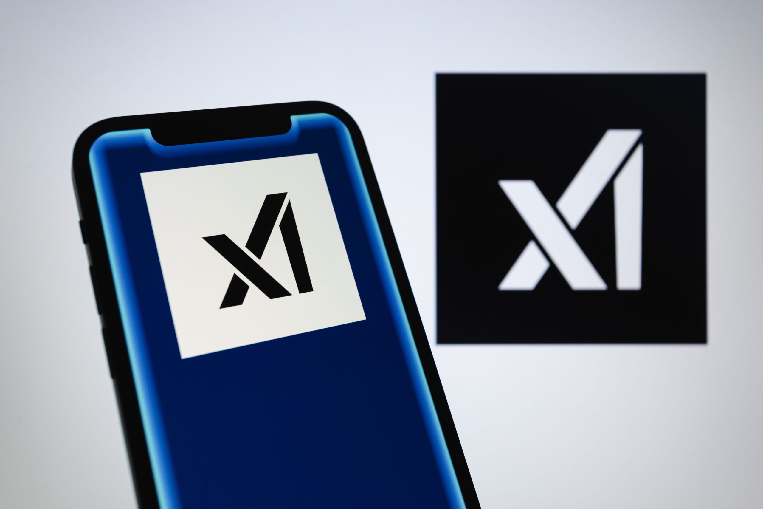 Logos von Elon Musks KI-Firma xAI auf einem Smartphone und einem Bildschirm. Bei xAI wurde der Chatbot Grok entwickelt, der unter anderem über die Online-Plattform X verfügbar ist.