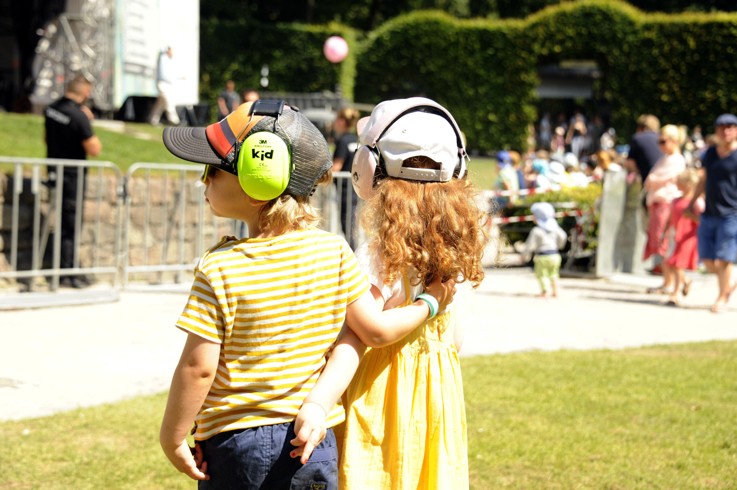 Zwei kleine Kinder stehen bei Sonnenschein Arm in Arm vor der Stadtpark-Bühne, beide mit Gehörschutz und Caps. Im Hintergrund weitere Menschen und die Bühne