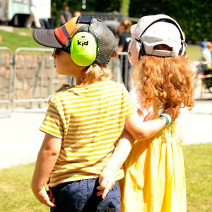 Zwei kleine Kinder stehen bei Sonnenschein Arm in Arm vor der Stadtpark-Bühne, beide mit Gehörschutz und Caps. Im Hintergrund weitere Menschen und die Bühne
