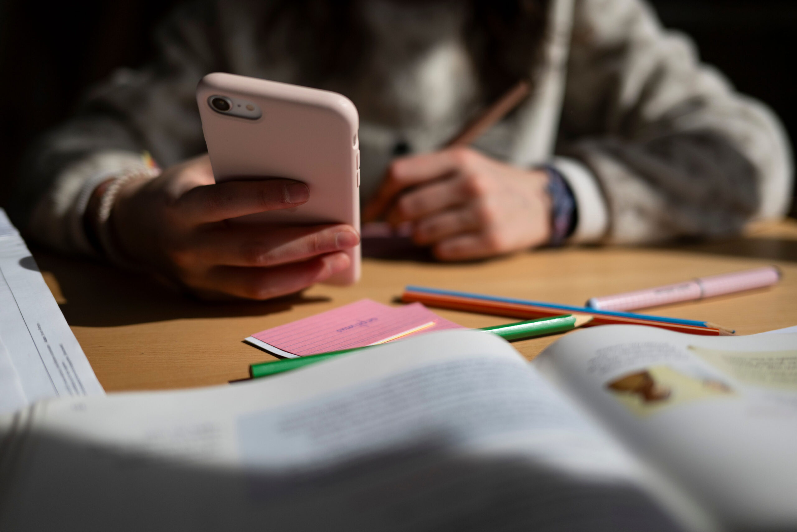 in Schulkind sitzt am Schreibtisch, bearbeitet Aufgaben mit einem Stift und hält dabei ein Smartphone in der Hand.