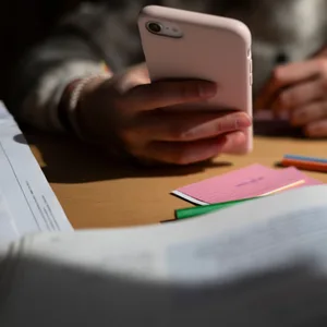 in Schulkind sitzt am Schreibtisch, bearbeitet Aufgaben mit einem Stift und hält dabei ein Smartphone in der Hand.