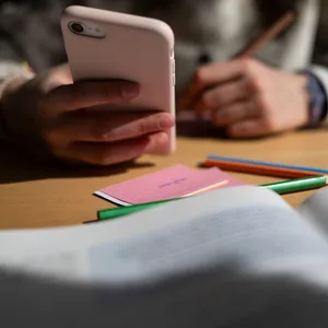 Ein Schulkind sitzt am Schreibtisch, bearbeitet Aufgaben mit einem Stift und hält dabei ein Smartphone in der Hand.
