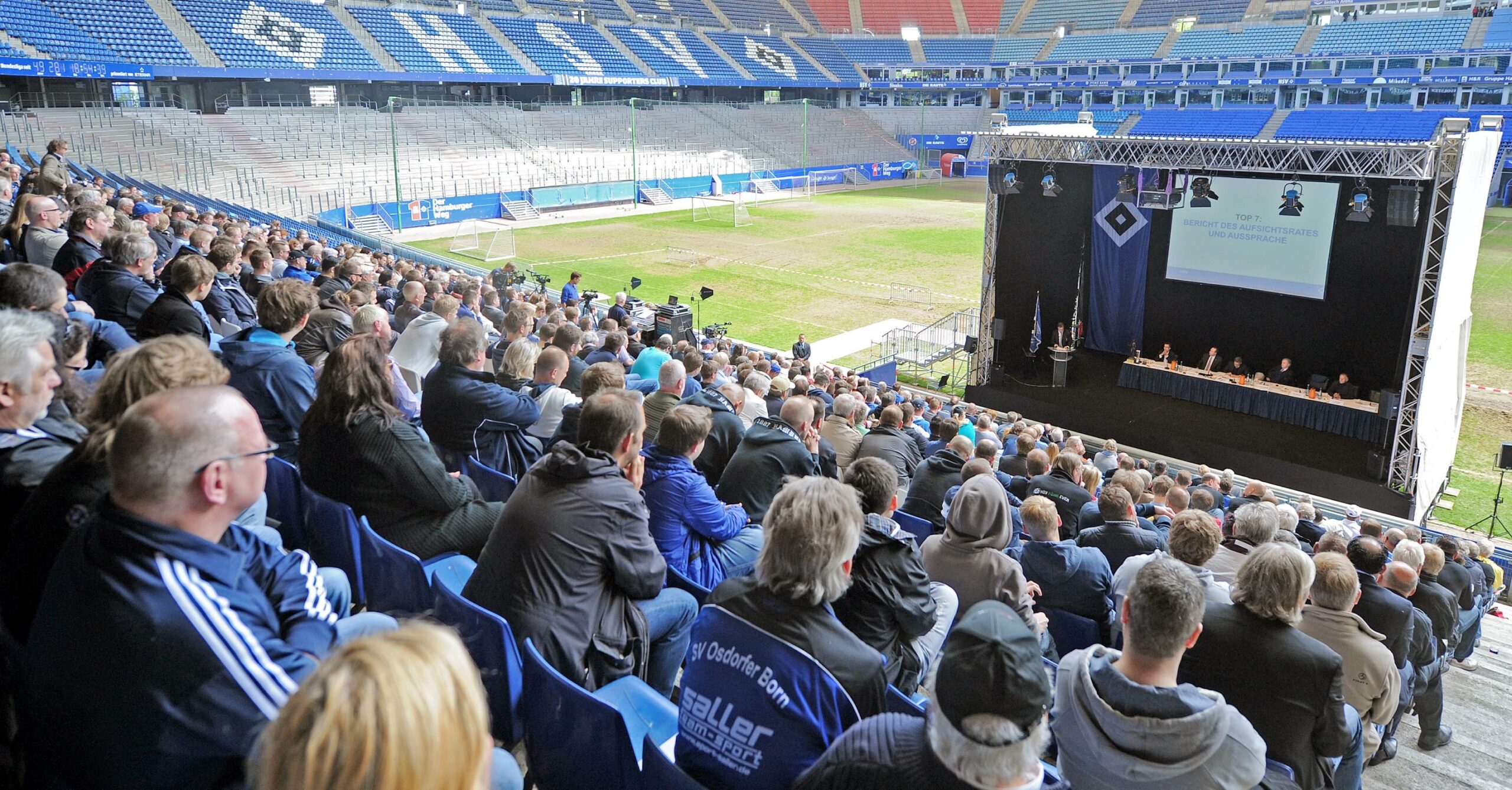 HSV-Mitglieder schauen von der Tribüne auf eine Bühne am Spielfeldrand des Volksparkstadions