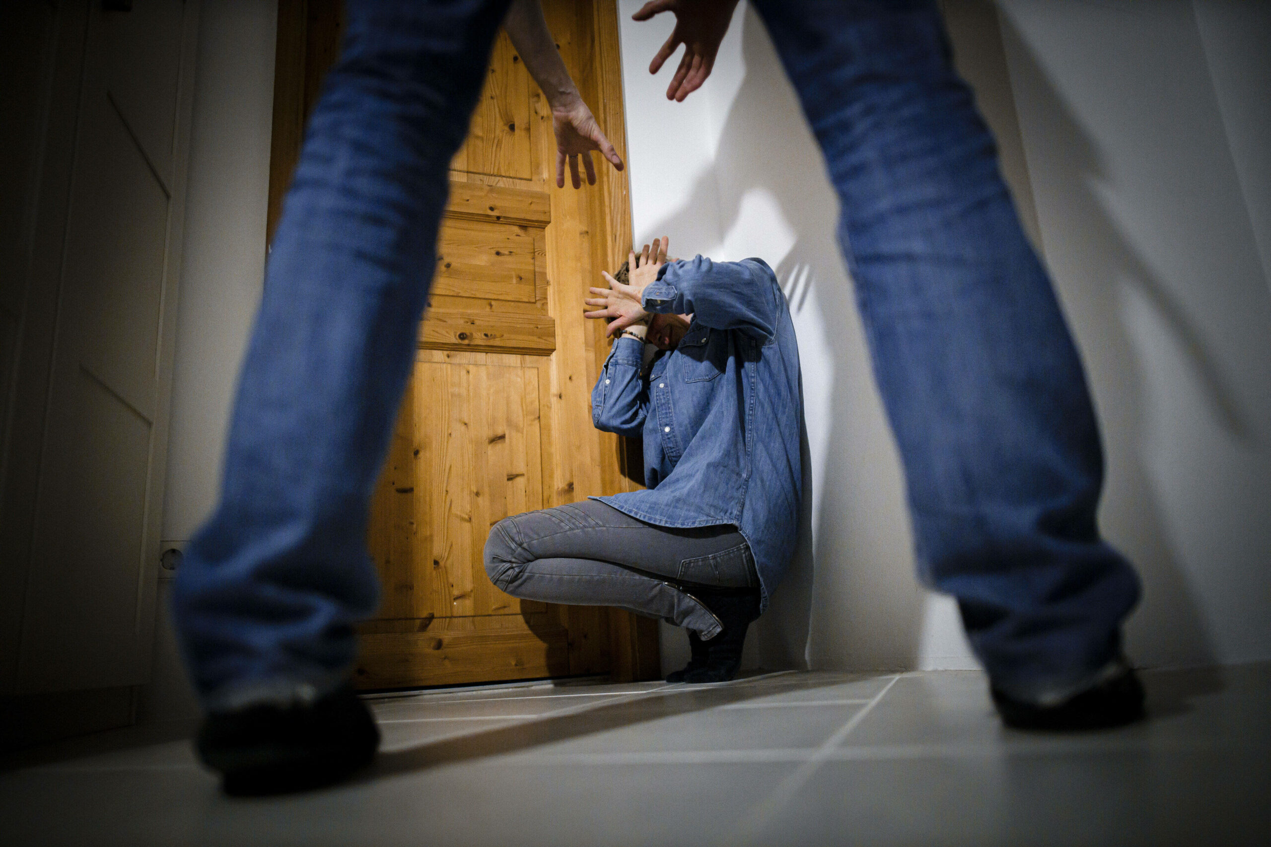Man schaut durch zwei Beine auf eine am Boden kauernde Person, die ihr Gesicht mit den Händen schützt.