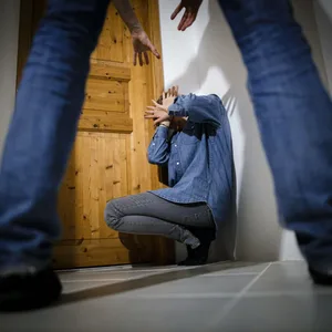 Man schaut durch zwei Beine auf eine am Boden kauernde Person, die ihr Gesicht mit den Händen schützt.