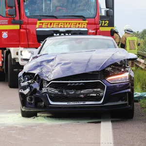 Ein zerbeulter schwarzer Audi steht vor einem Einsatzwagen der Feuerwehr.