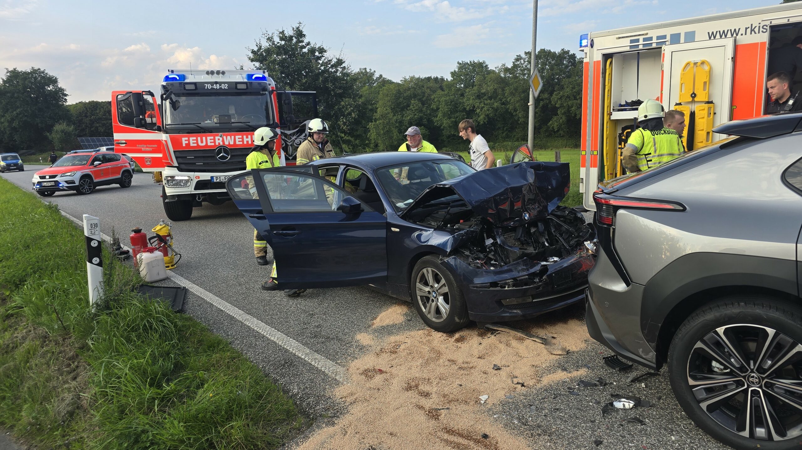 Ein schwarzes Auto, steht stark beschädigt hinter einem grauen Pkw. Zwei Feuerwehrautos stehen ebenfalls an der Straße.