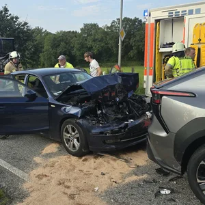 Ein schwarzes Auto, steht stark beschädigt hinter einem grauen Pkw. Zwei Feuerwehrautos stehen ebenfalls an der Straße.