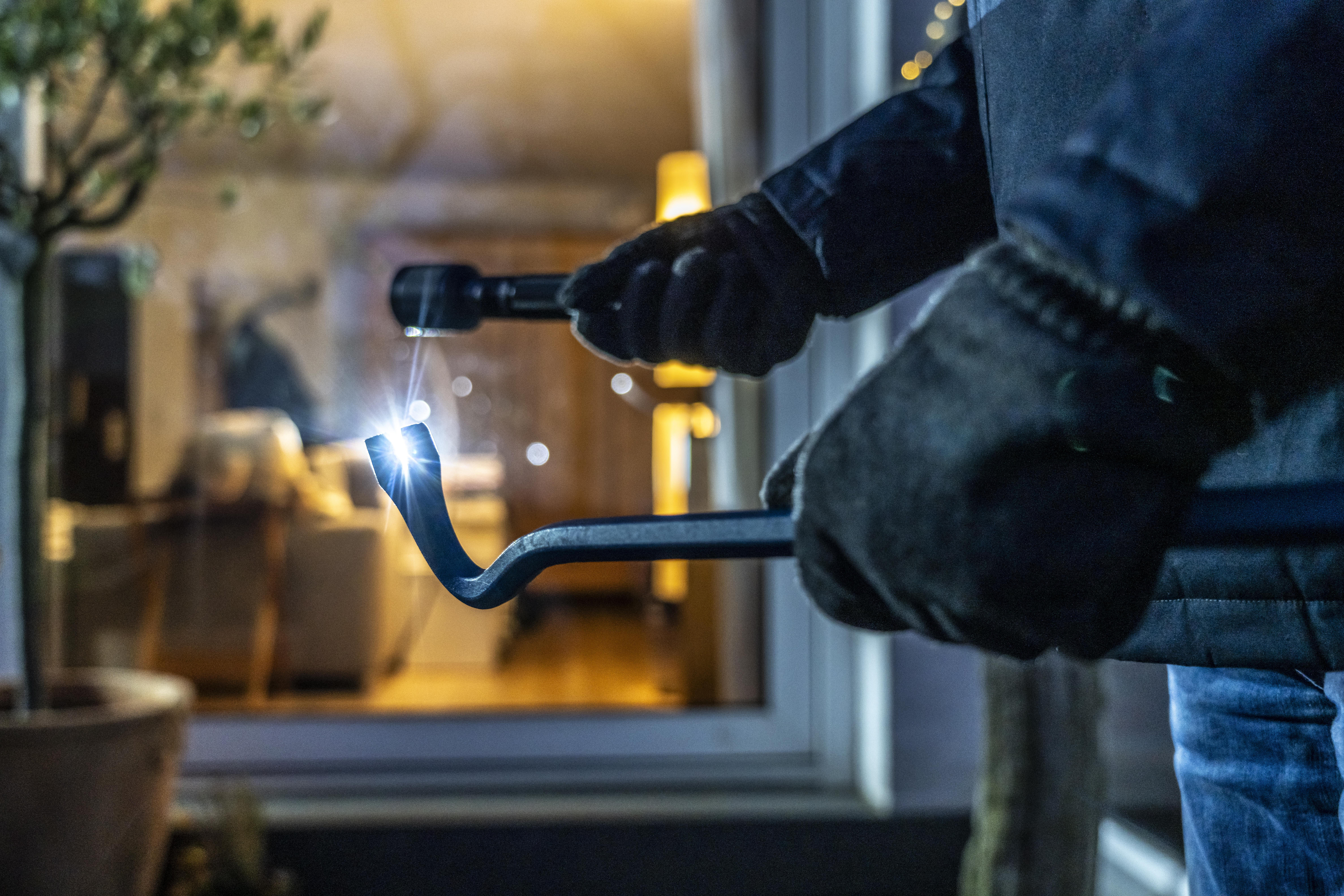 Ein Einbrecher steht mit Taschenlampe und Brecheisen vor einem Haus