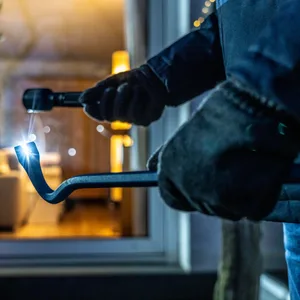 Ein Einbrecher steht mit Taschenlampe und Brecheisen vor einem Haus