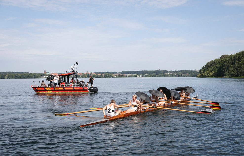 Schauspieler und professionelle Ruderer im Deutschland-Achter schützen sich bei Dreharbeiten auf dem Ratzeburger Küchensee zu dem Kinofilm „Adams Acht“ mit Schirmen vor der Sonne. 