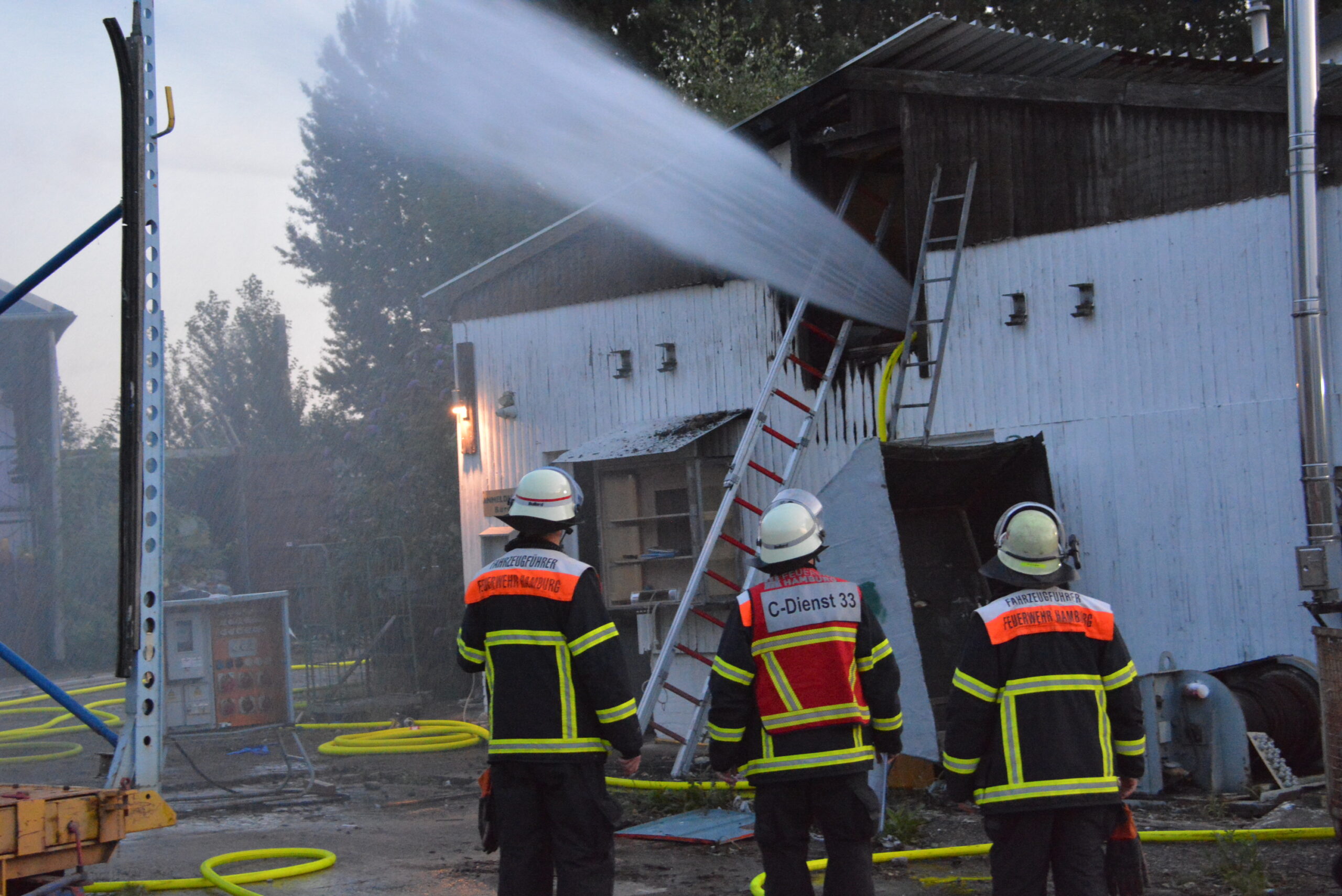 Drei Feuerwehrleute stehen vor dem Werftgebäude. Ein riesiger Wasserstrahl kommt aus dem oberen Geschoss.