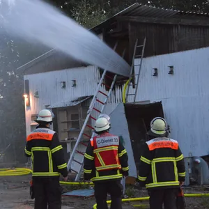 Drei Feuerwehrleute stehen vor dem Werftgebäude. Ein riesiger Wasserstrahl kommt aus dem oberen Geschoss.