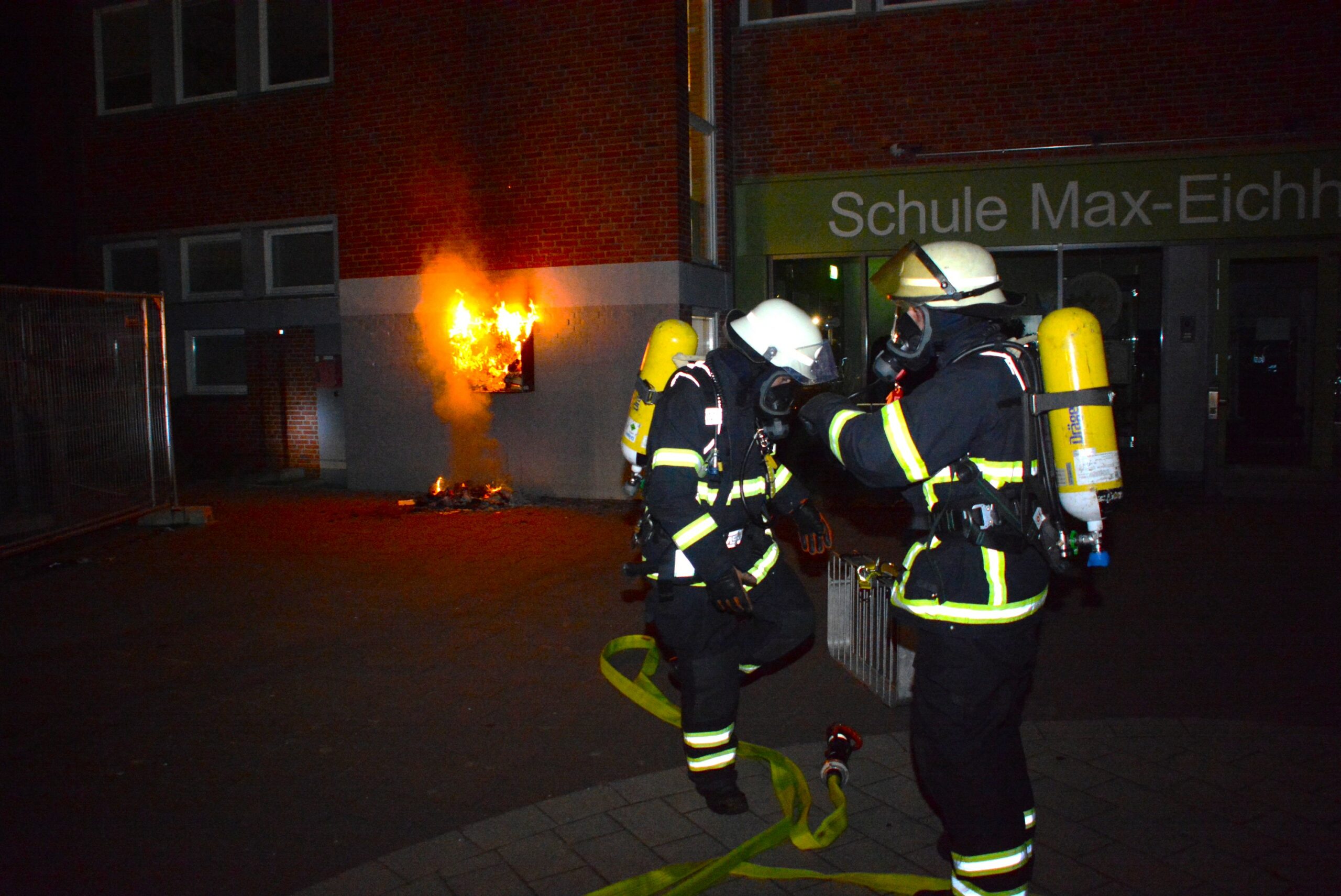 Einsatzkräfte der Feuerwehr stehen vor der Fassade der Grundschule, an der ein Bücherregal brennt.