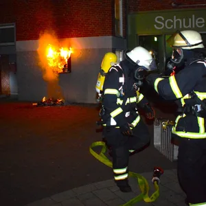 Einsatzkräfte der Feuerwehr stehen vor der Fassade der Grundschule, an der ein Bücherregal brennt.