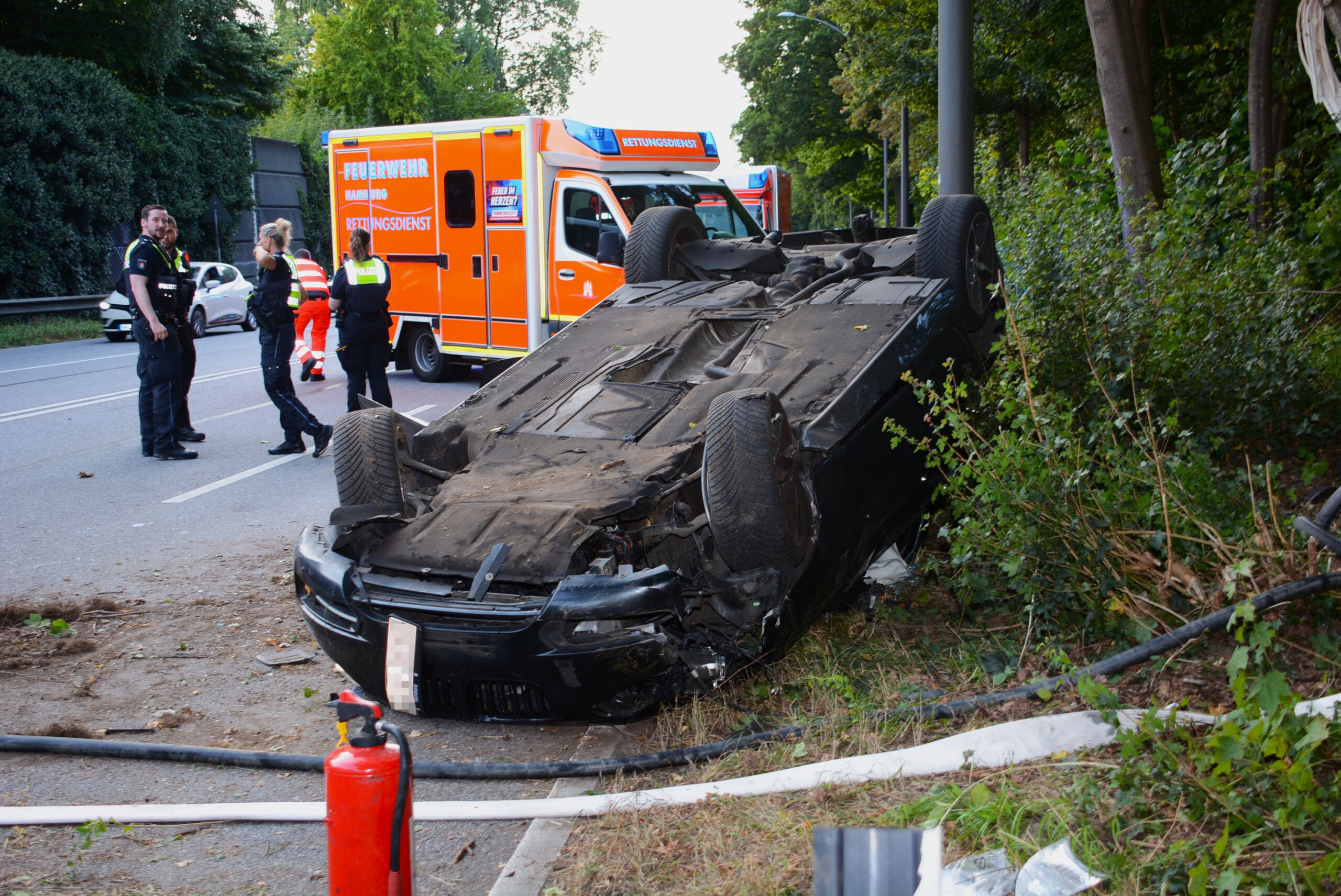 Ein schwarzes Auto liegt am Straßenrand auf dem Dach. Polizisten stehen links daneben.