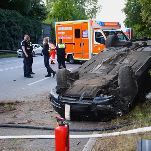 Ein schwarzes Auto liegt am Straßenrand auf dem Dach. Polizisten stehen links daneben.