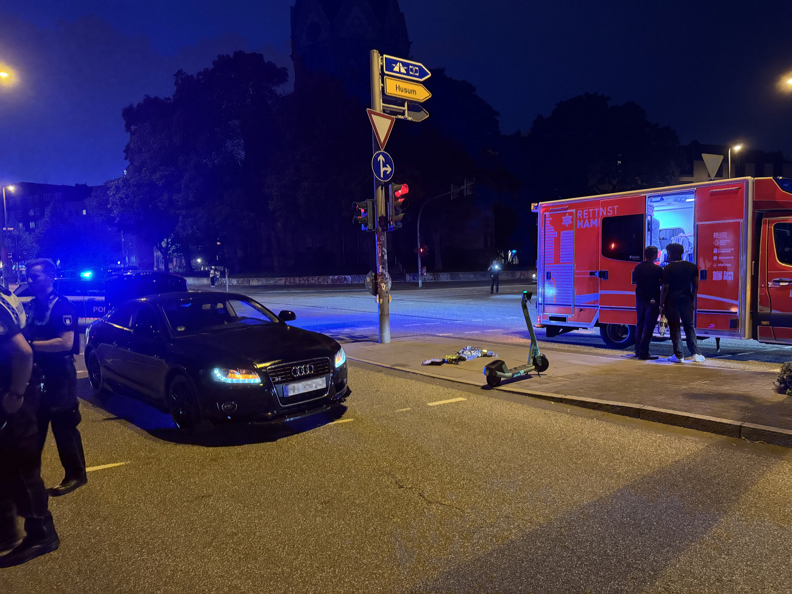 Ein schwarzer Audi steht an einer Ampel. Rechts von dem Auto steht der E-Scooter.