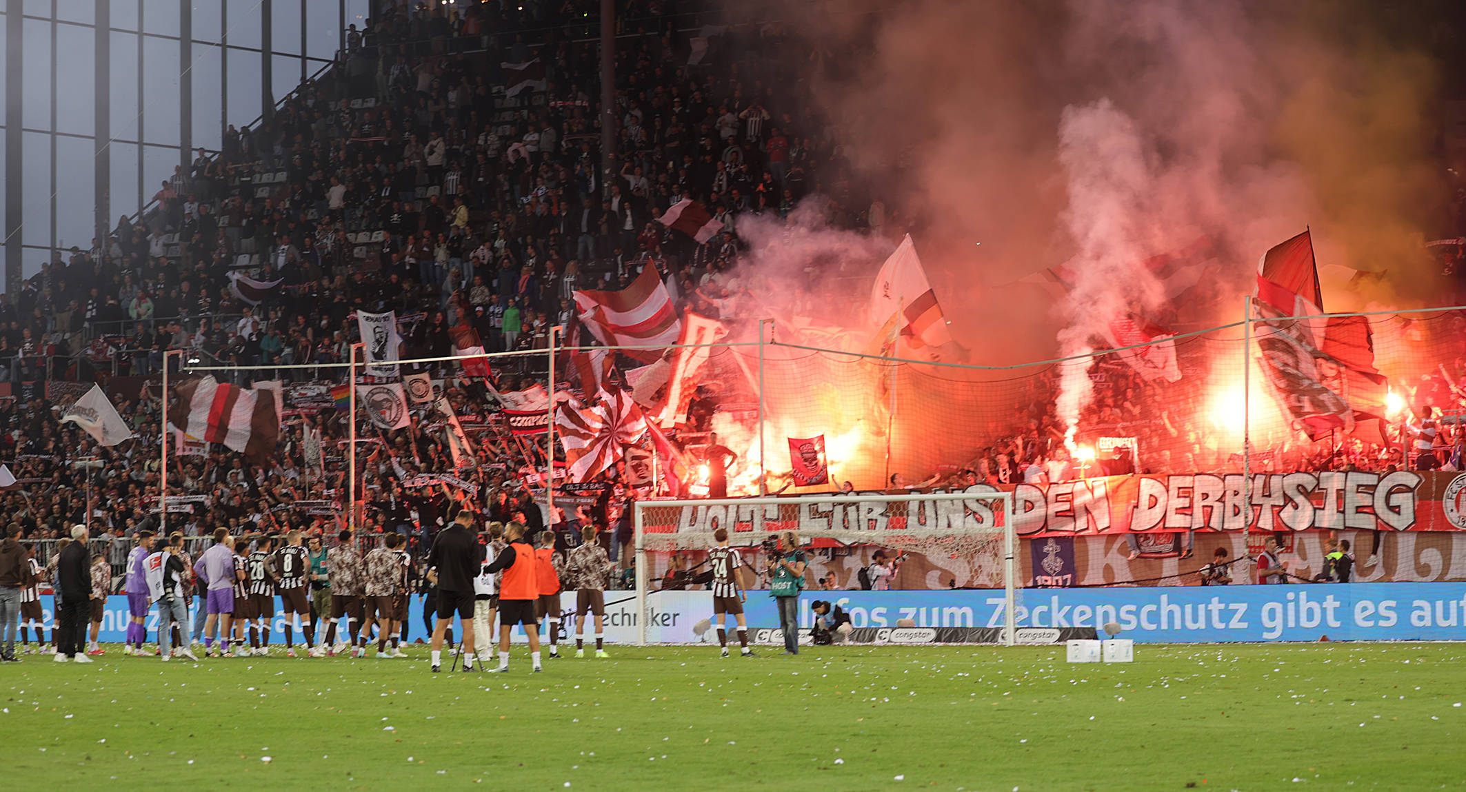 „Holt für uns den Derbysieg“ steht auf einem Banner im Fanblock, es brennt Pyrorechnik.