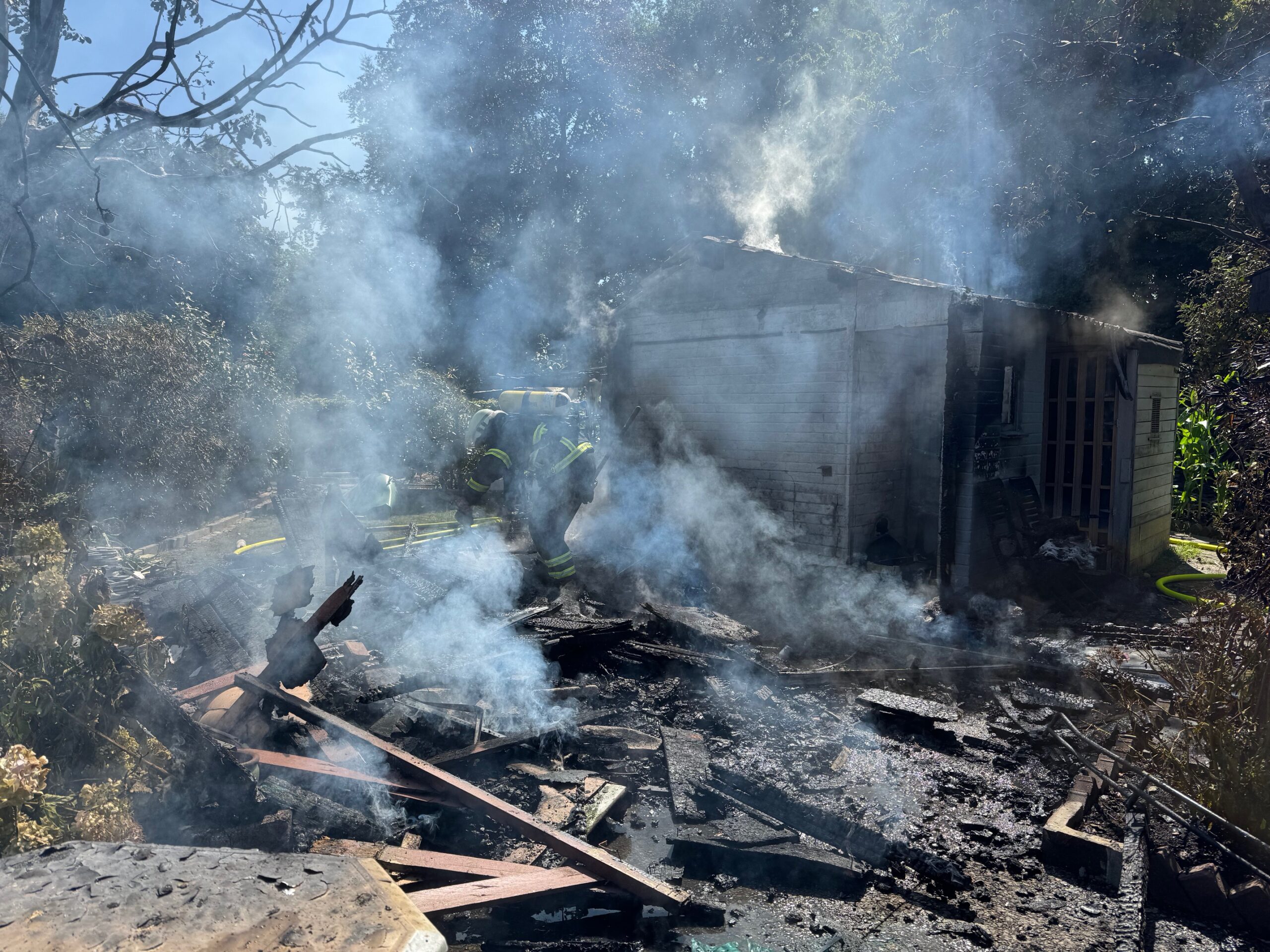 Feuerwehrkräfte stehen vor den rauchenden Überresten der abgebrannten Gartenlaube.