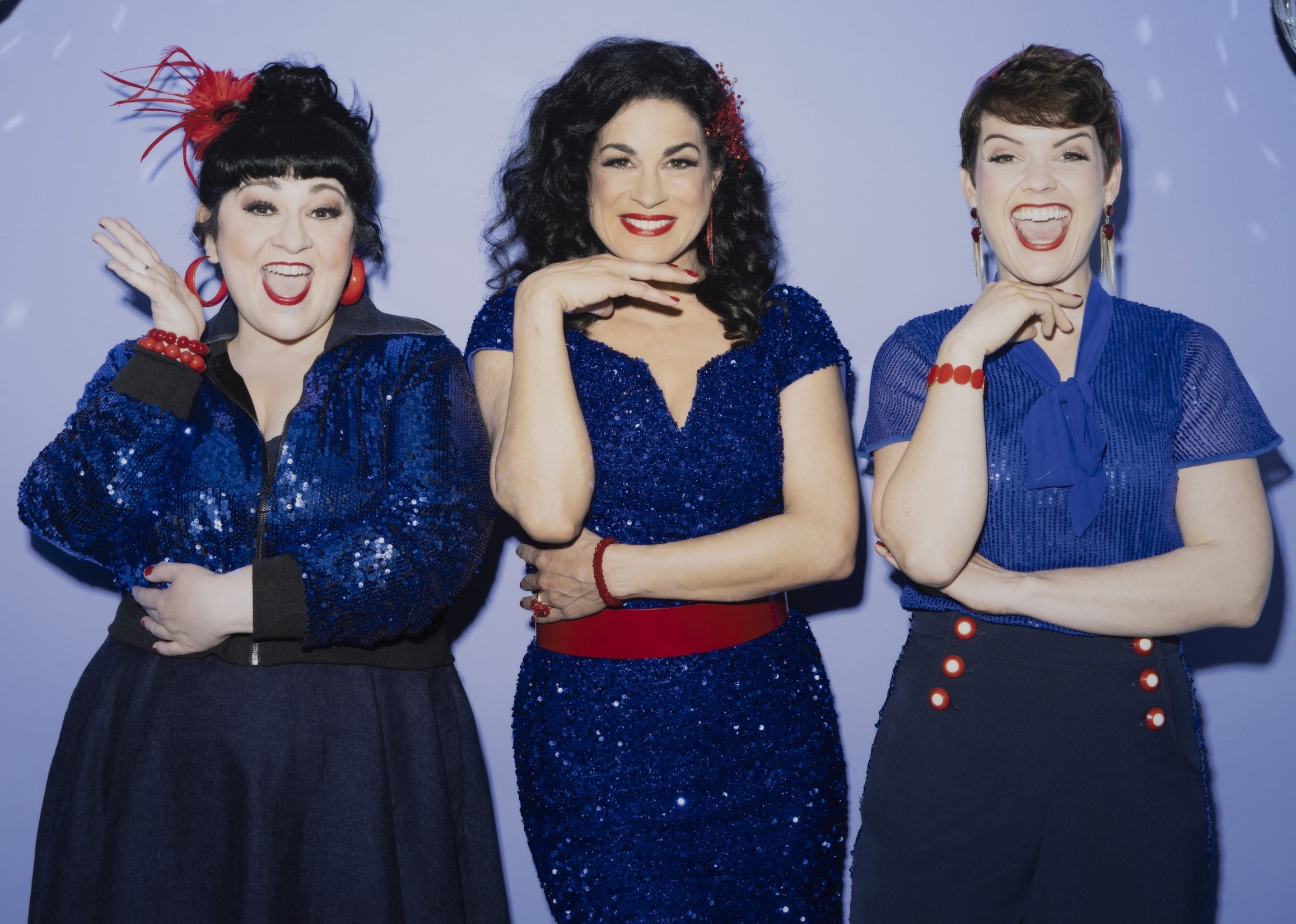 Drei Frauen in blauen Kleidern mit roten Details lachen in die Kamera