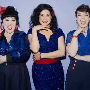 Drei Frauen in blauen Kleidern mit roten Details lachen in die Kamera