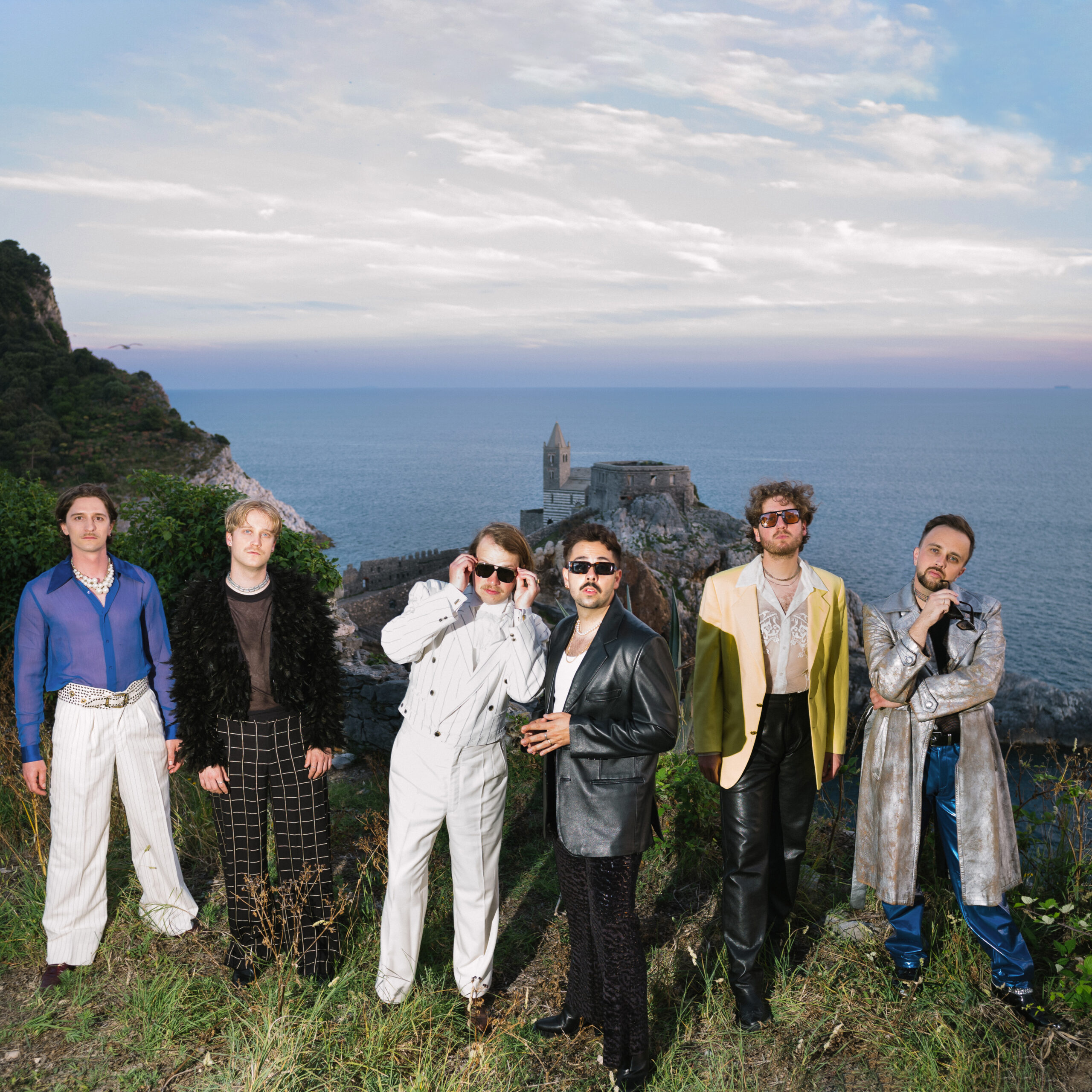 Sechs Männer am Rande einer Klippe, hinter ihnen das Meer und ein Gebäude auf einem Felsen