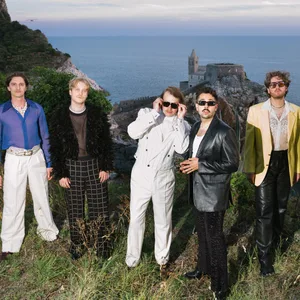 Sechs Männer am Rande einer Klippe, hinter ihnen das Meer und ein Gebäude auf einem Felsen