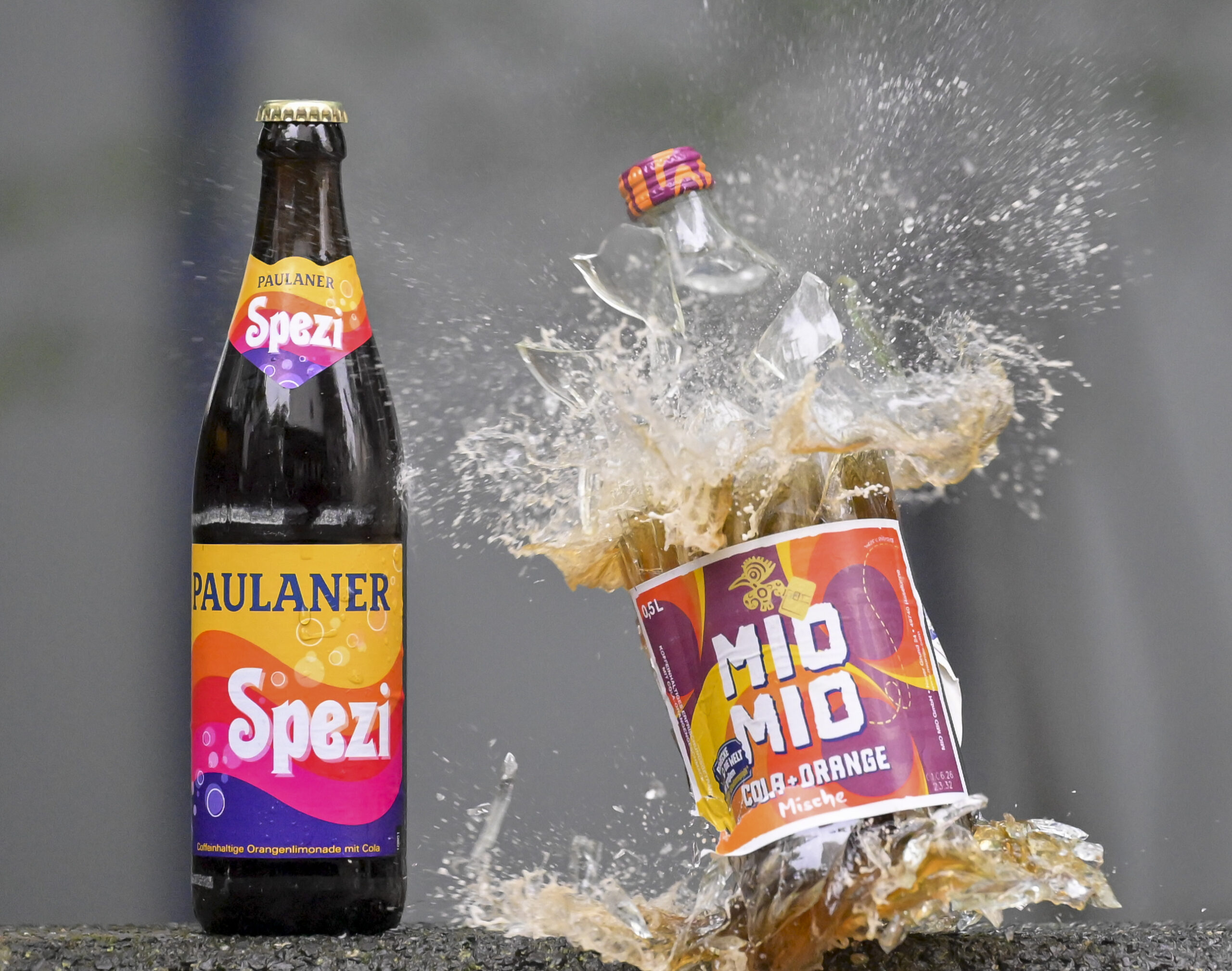 Eine Spezi-Flasche des Herstellers Paulaner (l) steht neben einer zerberstenden Flasche Mio-Mio-Cola-Orange-Mische des Herstellers Berentzen (r).