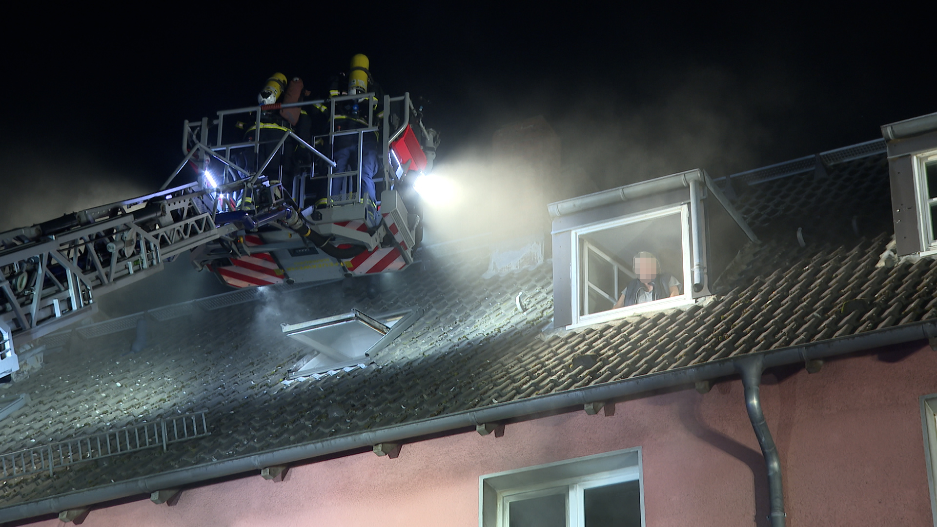 Die Feuerwehr steht mit einem Kran vor dem Fenster eines Mehrfamilienhauses. Im Fenster steht eine Person, das Gesicht ist gepixelt.