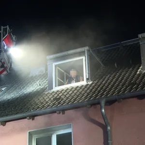 Die Feuerwehr steht mit einem Kran vor dem Fenster eines Mehrfamilienhauses. Im Fenster steht eine Person, das Gesicht ist gepixelt.