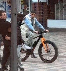 Ein Mann in blauem Pulli und beige/weißer Weste auf einem Fahrrad.