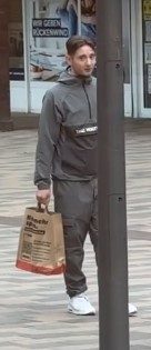 Ein junger Mann in grauer Hose und grauer Jacke geht mit einer Einkaufstüte den Weg entlang.
