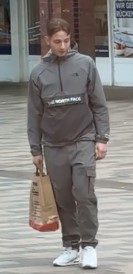 Ein junger Mann in grauer Hose und grauer Jacke geht mit einer Einkaufstüte den Weg entlang.