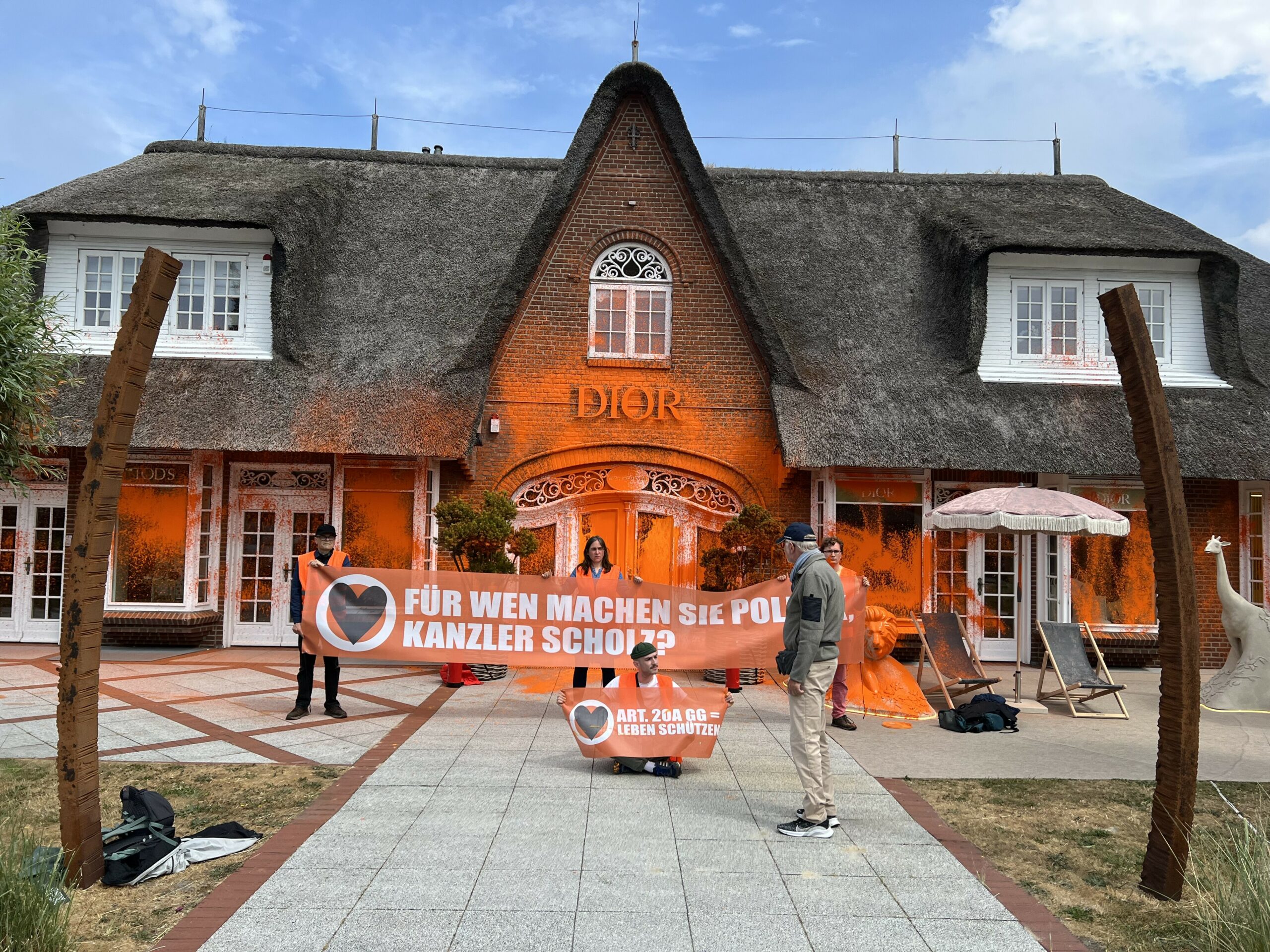 Demonstranten der Letzten Generation stehen und sitzen vor einem Luxusgeschäft auf Sylt. Das Geschäft ist mit oranger Farbe besprüht.
