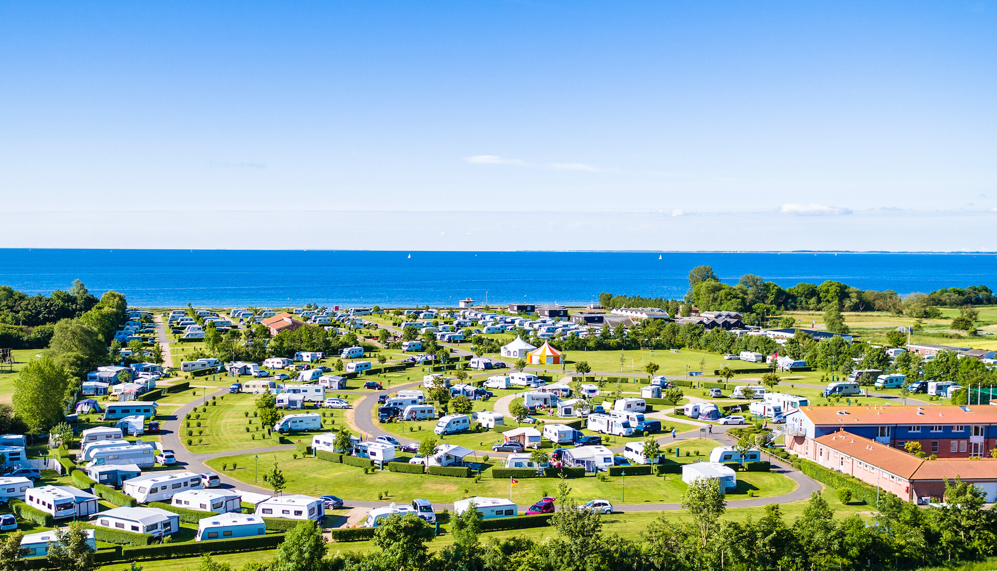 Das „Insel-Camp“ auf Fehmarn. Im Hintergrund ist blauer Himmel und blaues Meer. Man sieht viele weiße Wohnwagen/ Wohnmobile.