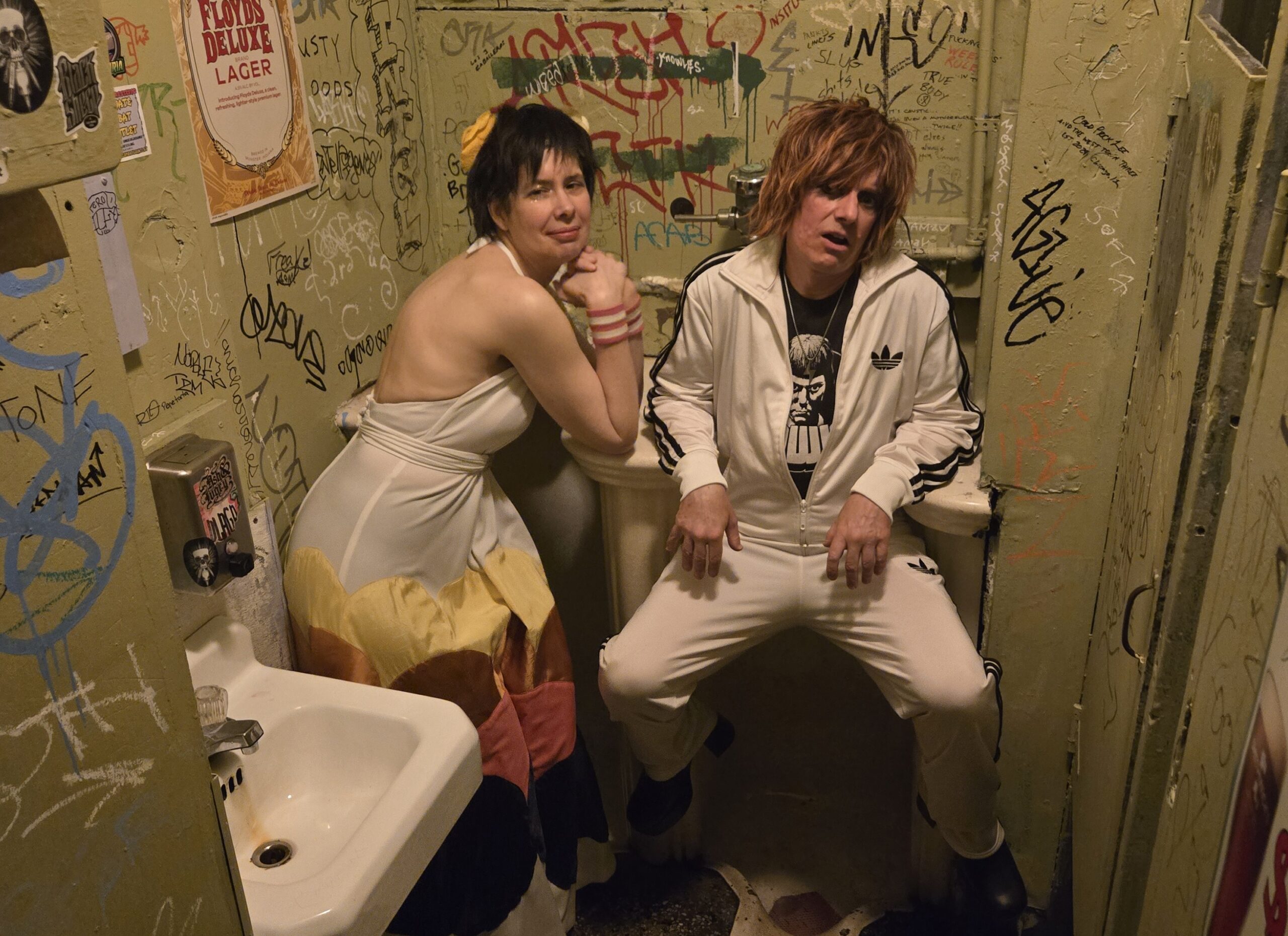 Ein Mann und eine Frau, beide tragen weiße Kleidung, sitzen auf einer Toilette, Aufkleber und Schriftzüge an den Wänden