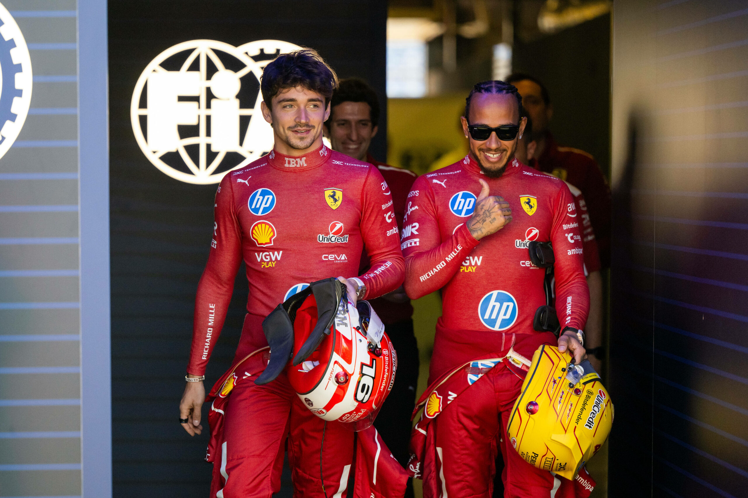 Lewis Hamilton neben Charles Leclerc, beide in Rennkleidung und mit Helm in der Hand.