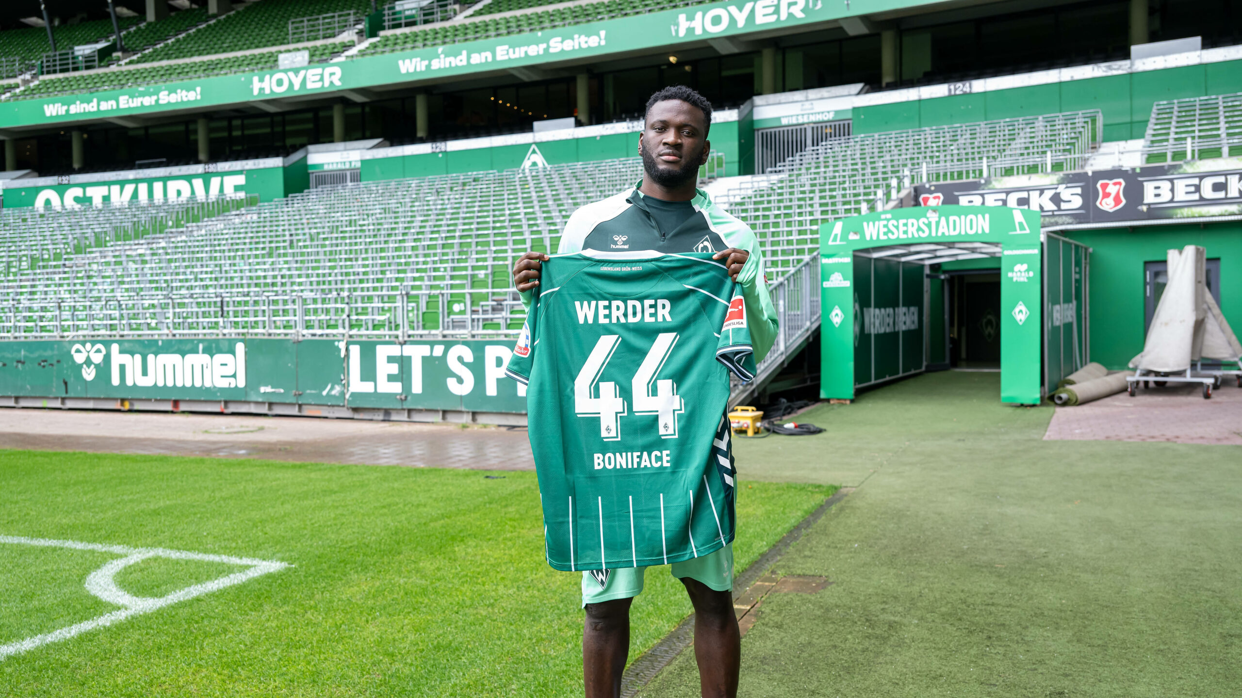 Boniface vor der leeren Ostkurve im Bremer Weserstadion. Er hält sein Trikot mit der 44 hoch.