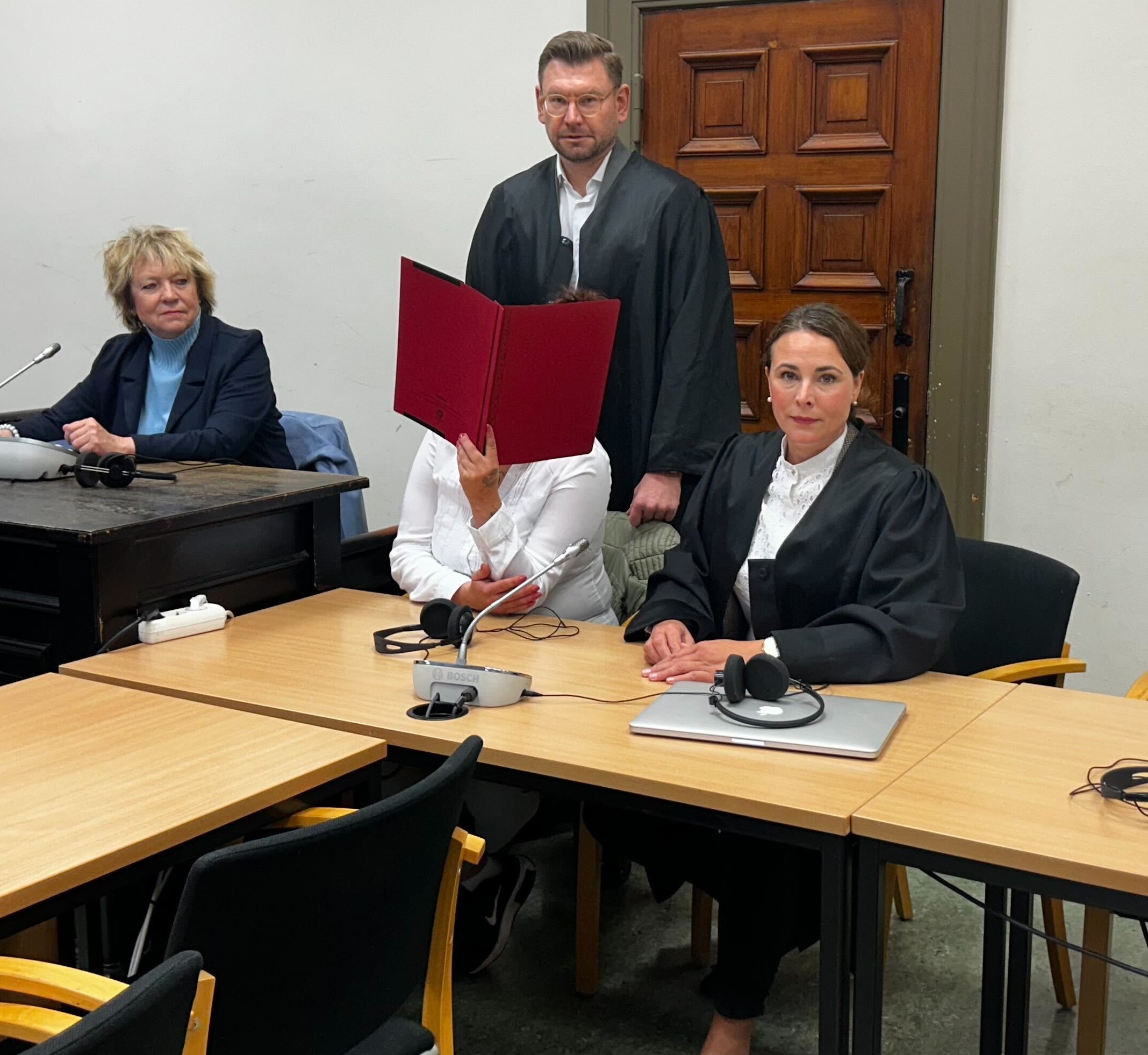 Die Angeklagte verdeckt ihr Gesicht mit einer Mappe. Ein Verteidiger steht hinter ihr. Die andere Verteidigerin sitzt rechts von ihr.