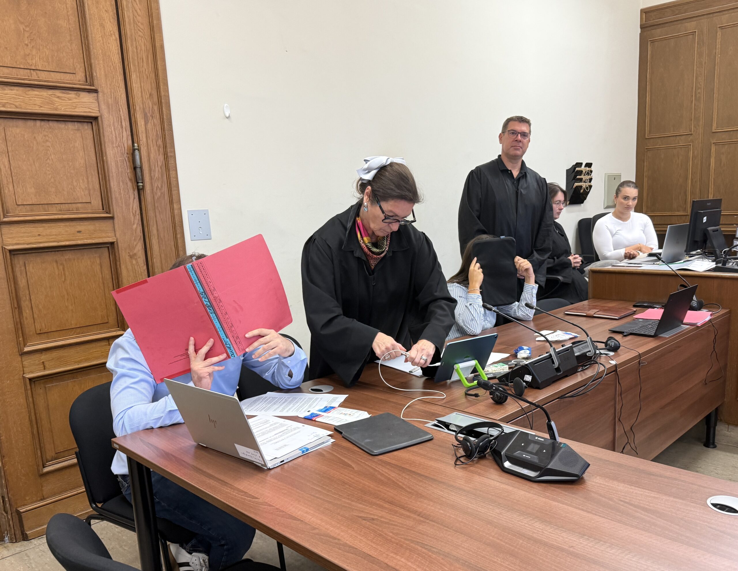 Peter O. hält sich eine rote Mappe vor das Gesicht. Seine Ehefrau Olga O. hält sich eine schwarze Laptop-Tasche vor ihr Gesicht.