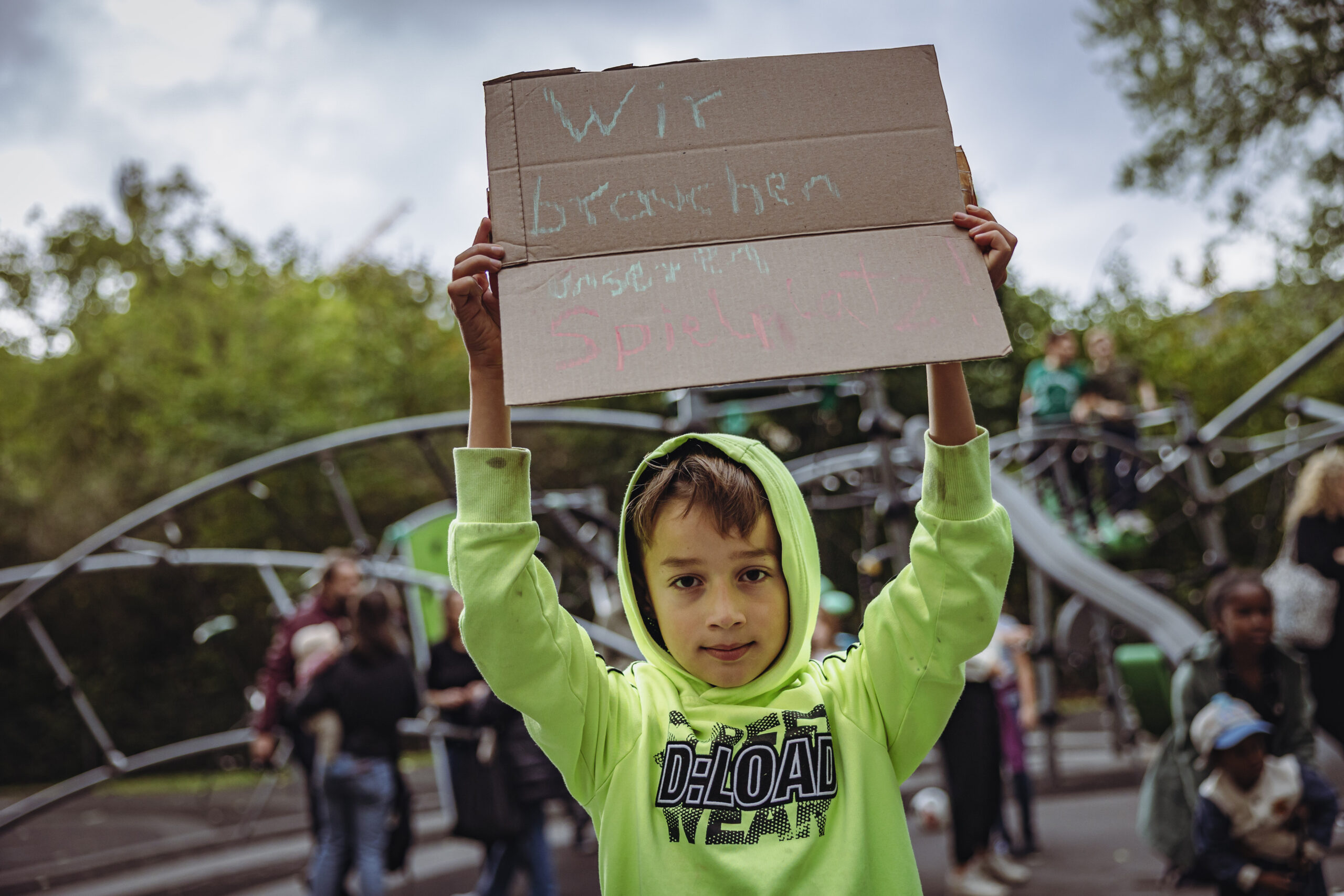 Ein Junge hält ein Schild in die Höhe. Darauf steht: „Wir brauchen unseren Spielplatz!“
