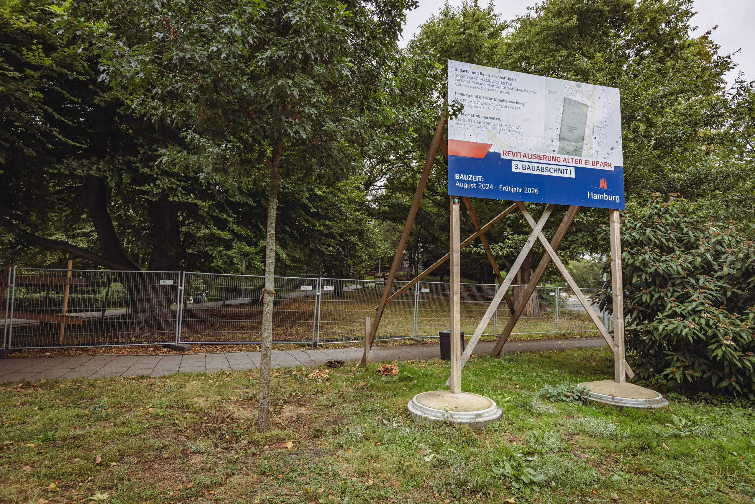 Ein Schild mit der Aufschrift „Bauzeit August 2024 bis Frühjahr 2026“ vor dem Bauzaun im Alten Elbpark.