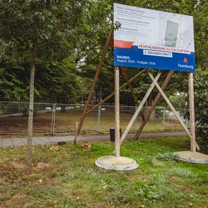 Ein Schild mit der Aufschrift „Bauzeit August 2024 bis Frühjahr 2026“ vor dem Bauzaun im Alten Elbpark.